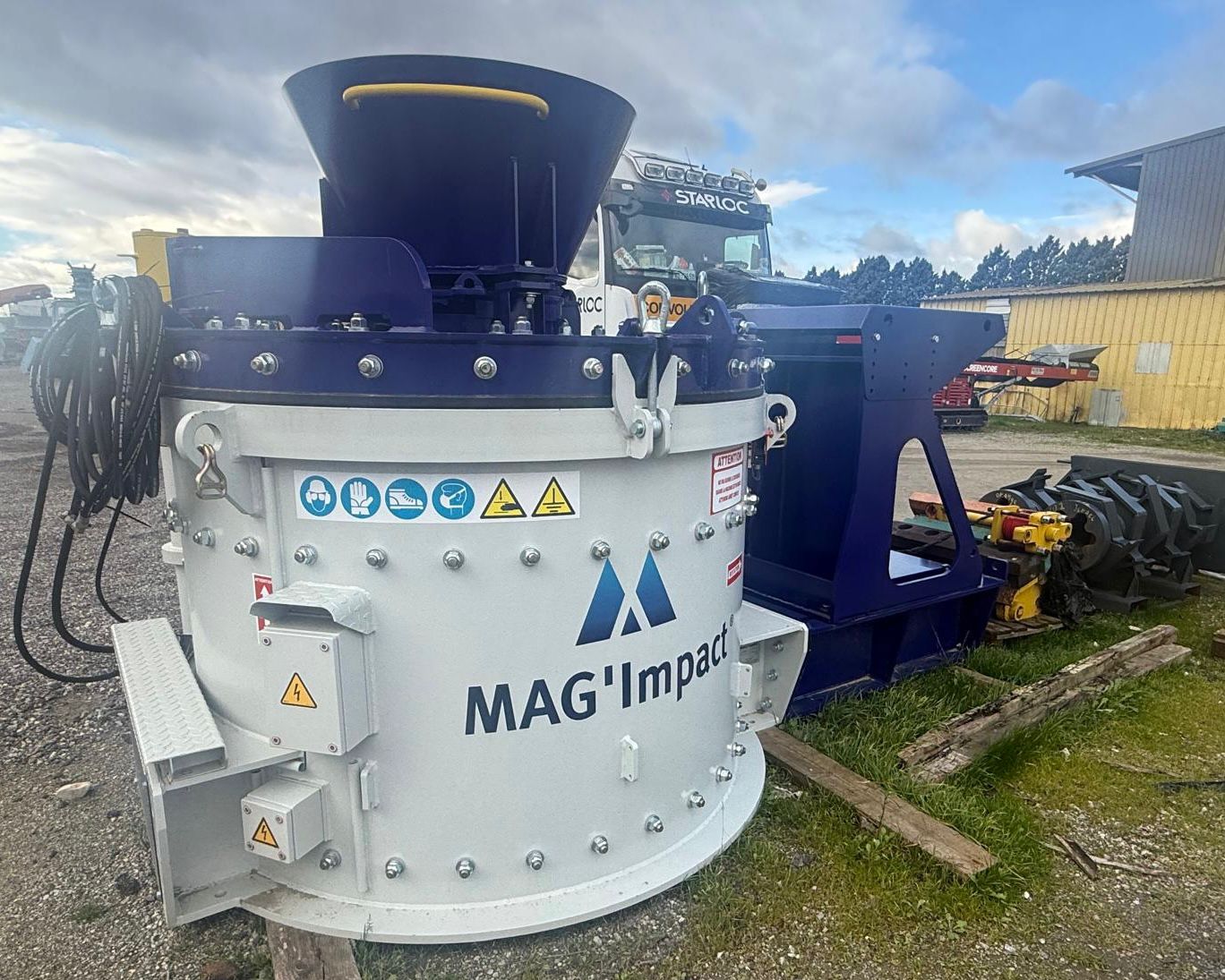 Maquinaria industrial de trituración MAG'Impact de color blanco y azul marino, con calcomanías de seguridad y componentes de acero, situada en un patio exterior con un camión de transporte al fondo.